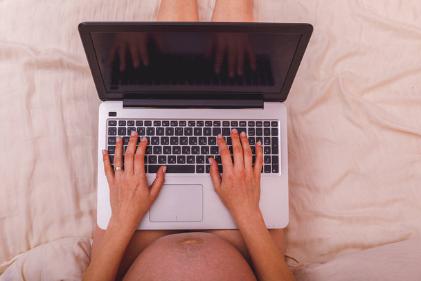 Beautiful pregnant young woman working on a laptop sitting on the bed in the bedroom. Concept of female business, shopping, home office, quarantine, coronavirus covid 19 ncov. Stay at home concept. - Фото, изображение