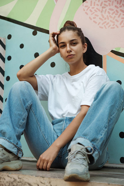 A beautiful young brunette in wide blue jeans and a white loose t-shirt is sitting on a bright multi-colored background. Beautiful fashionable young woman - Φωτογραφία, εικόνα