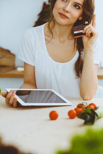Young woman looking for a new recipe for cooking in a kitchen. Housewife is making online shopping by tablet computer and credit card - Фото, зображення