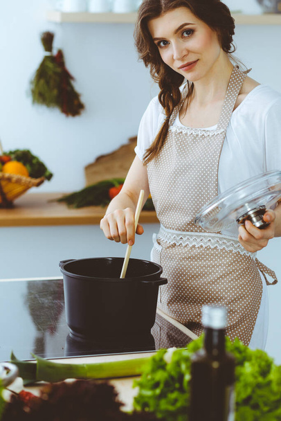 Young brunette woman cooking soup in kitchen. Housewife holding wooden spoon in her hand. Food and health concept - Photo, Image