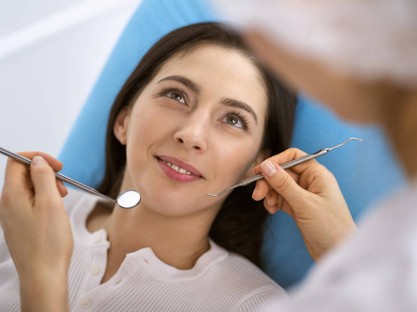 Smiling brunette woman being examined by dentist at dental clinic. Hands of a doctor holding dental instruments near patients mouth. Healthy teeth and medicine concept - Foto, afbeelding