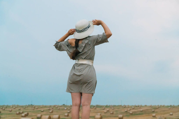 Beautiful girl in a hat looks into the sky in the middle of a wheat field with bales in summer evening - Fotografie, Obrázek