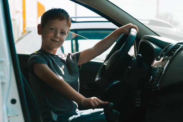 Little red-haired boy sits in front of the car holding steering wheel. Boy looking at the camera and smiling - Foto, immagini