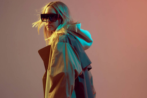 Stylish young girl, cool posing in a hoodie, sunglasses and cloak with developing hair, on a neon background. - Φωτογραφία, εικόνα
