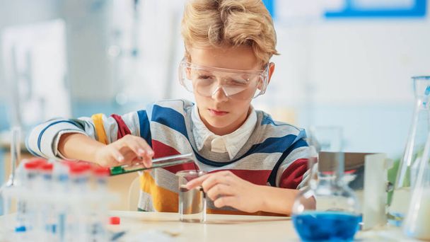 Elementary School Science or Chemistry Classroom: Smart Little Boy wearing Safety Glasses Mixes Chemicals in Beakers - Photo, Image