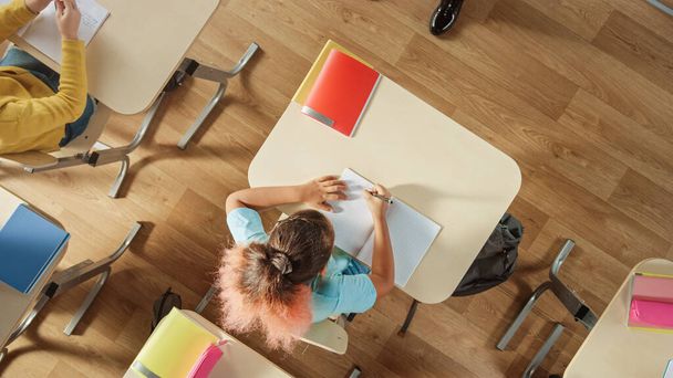 Top View Shot of Elementary School Classroom: Girl Sitting at the School Desk Working on Assignments in Exercise Notebooks. - Fotoğraf, Görsel