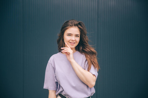 Young stylish trendy woman isolated over grey blue background. Cheerful positive girl in violet shirt and white pants holding hand close to chin and smile. Posing alone with happiness. - Photo, Image
