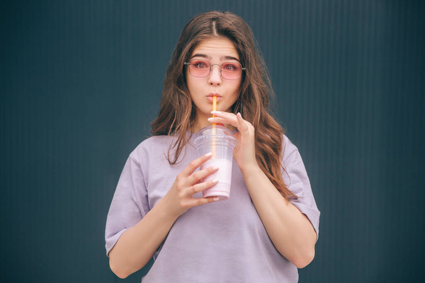 Young stylish trendy woman isolated over grey blue background. Emotional girl drinking milk cocktail and look straight on camera through glasses. Facial expression. Stand alone at blue grey wall. - Photo, Image