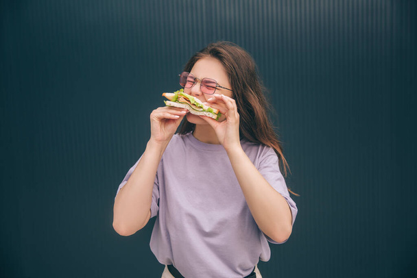 Jonge stijlvolle trendy vrouw geïsoleerd over grijs blauwe achtergrond. Hongerig meisje die een vers broodje bijt. Een vrouw die buiten aan de muur ontbijt of luncht. Alleen eten. - Foto, afbeelding