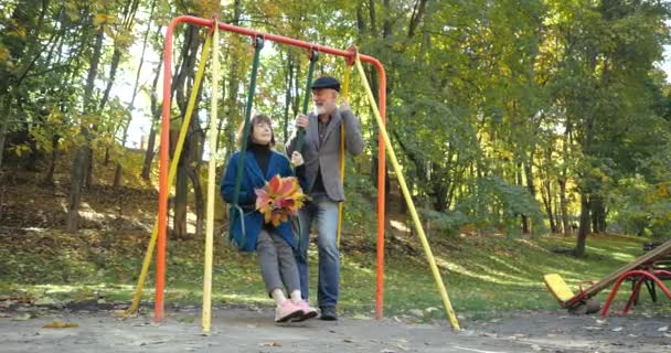 Sakallı koca, karısı elinde bir buket sonbahar yaprağı tutarken sallanan sandalyeyi sallıyor. Şımarık, aktif, yaşlı çift şehir parkında eğleniyor. Swnior emekliler ailesi dışarıda eğleniyor.. - Video, Çekim