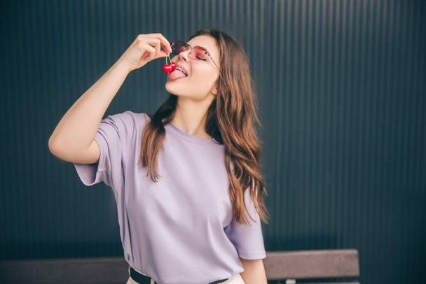 Young stylish trendy woman isolated over grey blue background. Happy girl holding two cherrie berries up in air and ready to eat them. Delicious healthy dessert. - Fotografie, Obrázek