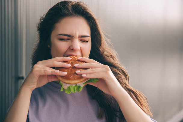 Joven mujer con estilo de moda aislado sobre fondo gris azul. Chica sosteniendo hamburguesa con dos manos y mordiendo parte de ella. Sabrosa comida deliciosa durante el almuerzo
. - Foto, imagen