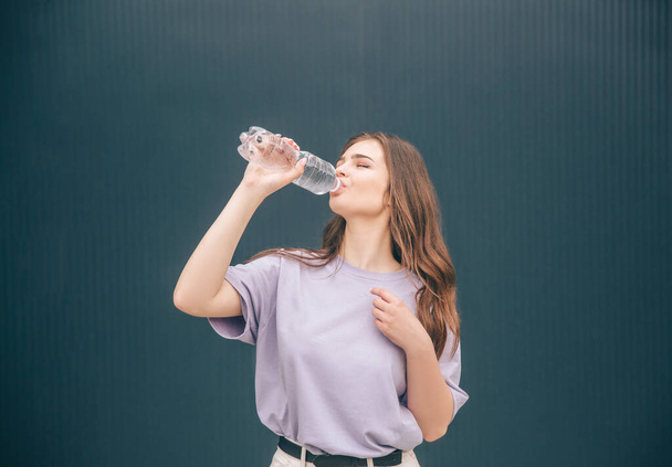 Młoda stylowa, modna kobieta odizolowana na szarym tle. Dziewczyna pije przezroczystą, smaczną wodę z przezroczystej plastikowej butelki. Nawodnienie ciała. - Zdjęcie, obraz