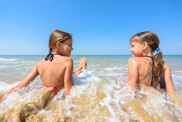 Kinder sitzen im Wasser an einem Sandstrand und schauen einander fröhlich an - Foto, Bild