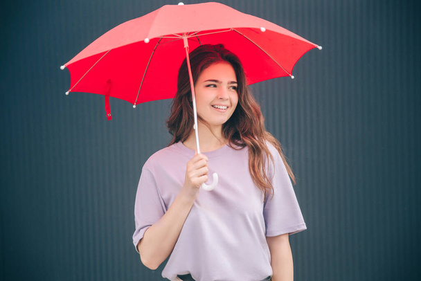 Young stylish trendy woman isolated over grey blue background. Positive cheerful girl standing under red umbrella and look right. Smiling cheerfully. Alone at dark wall. Rain cover and protection. - Photo, Image