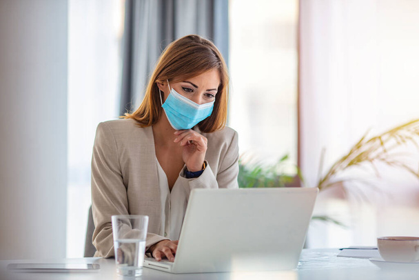 Female employee wearing medical face mask while working in the business office during covid-19 pandemic. Businesswoman wearing face mask while analzying reports in the office.  - Foto, Imagem