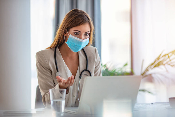 Female doctor counseling, helping patient online, medic using laptop for video call. Online doctor with a protective mask tutoring over the internet. Mid adult female doctor working in office - Photo, Image