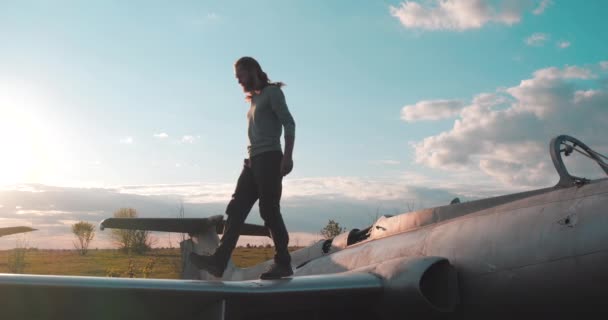 Bearded tourist walks on the wing of broken abandoned fighter plane at airfield in field and looks hopefully at the bright sun in slow motion. Active man explores cemetery of combat aircraft bombers. - Footage, Video