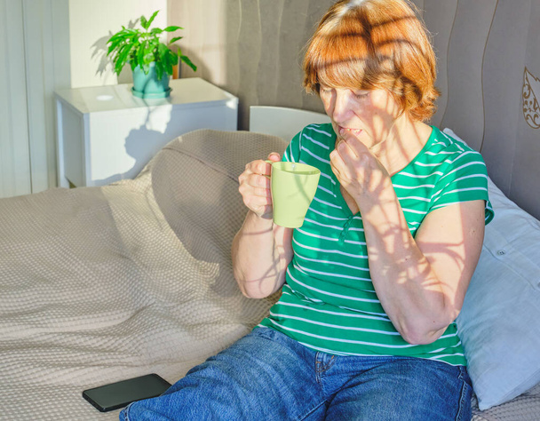 Senior woman feeling pain sitting on bed at home in casual clothes. In one hand she holds a pill, about to take it, in the other a cup of water. - Photo, Image
