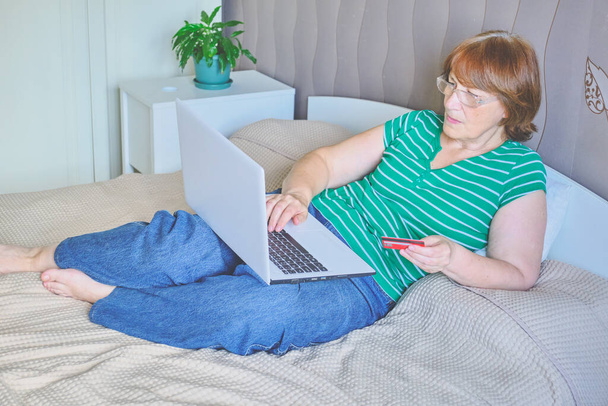 Vanhempi nainen, joka käyttää läppäriä kotonaan. Onnellinen eläkkeellä henkilö ostoksia verkossa sängyllä. Elämäntapa. Kopioi tila - Valokuva, kuva