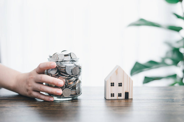 Hand holding jar/bottle glass bank that contain full of coins next to woodden house model and tree house decoration background. Concept of financial money saving/ house or real estate investment. - 写真・画像