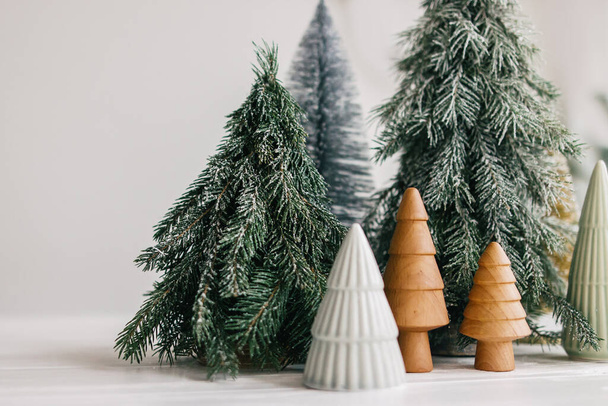 Christmas little trees on white background. Winter forest, festive modern decor. Happy holidays. Miniature ceramic,wooden, snowy and handmade pine trees. Season greetings. Merry Christmas. - Фото, зображення