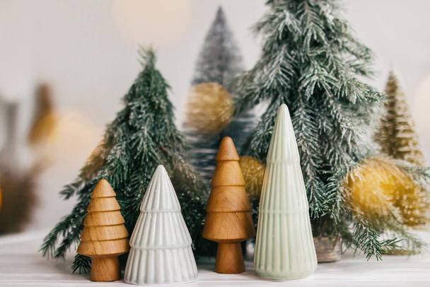 メリークリスマス。白い背景にクリスマスの小さな木や金色のライトボケ。冬の魔法の森、お祝いのモダンな装飾。ミニチュアセラミック、木製、雪と手作りの松の木. - 写真・画像