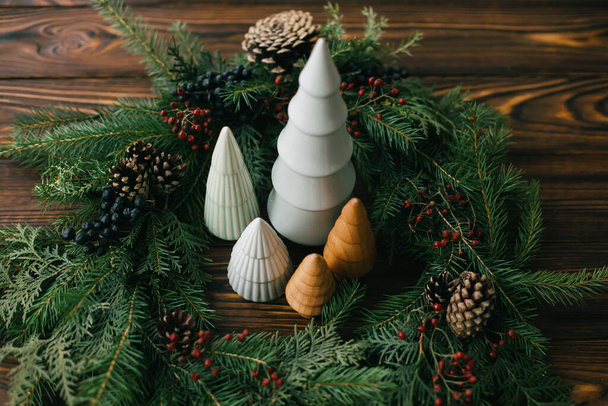 Weihnachtsbäume in stilvollem rustikalem Weihnachtskranz auf dunklem Holztisch. Festliches, modernes Dekor, keine Verschwendung. Frohe Feiertage. Miniatur-Kiefern und moderner Kranz mit Beeren und Tannenzapfen - Foto, Bild