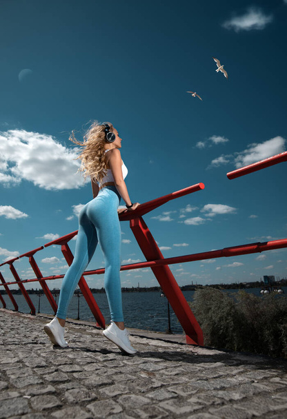 Beautiful fitness girl posing on the shore cliff of the river background in sexy blue leggings and earphones in the rays of the rising sun and the seagulls flying in the sky. - Valokuva, kuva