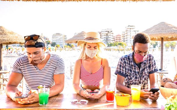 Multiracial friends with closed face masks using tracking app with mobile smartphones - Bored young milenial people at beach cocktail bar - New normal lifestyle concept - Warm vivid backlight filter - Photo, Image