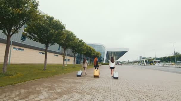 旅に出る3人の若い幸せな女の子に同行する男。女性は空港に手荷物を持って歩く。男は手を振って - 映像、動画