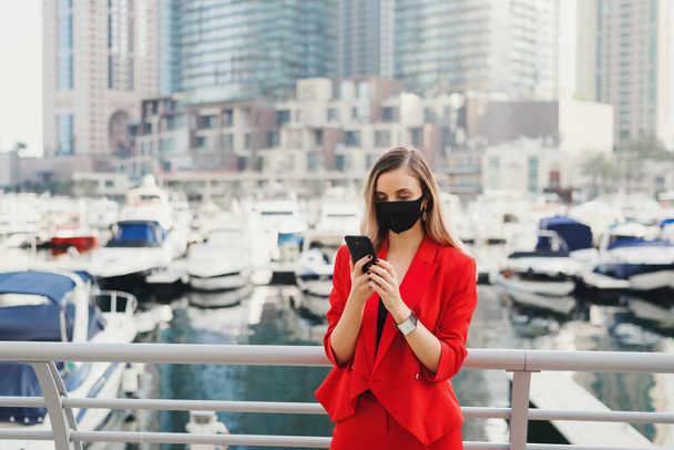 Młoda przedsiębiorczyni stojąca w centrum biznesowym miasta przed luksusowymi jachtami i drapaczami chmur i patrząca na ekran smartfona. Stylowa kobieta w czerwonym garniturze i masce ochronnej twarzy - Zdjęcie, obraz