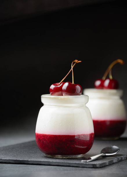 Dessert in jar with berries no baking. Italian panna cotta with sweet cherry on black slate board, dark concrete background. - Valokuva, kuva
