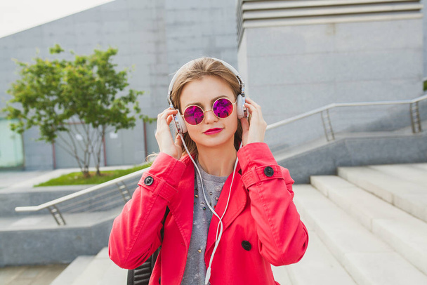 young hipster woman in pink coat, jeans in street with coffee listening to music on headphones, big city urban style trend, wearing sunglasses - Photo, image
