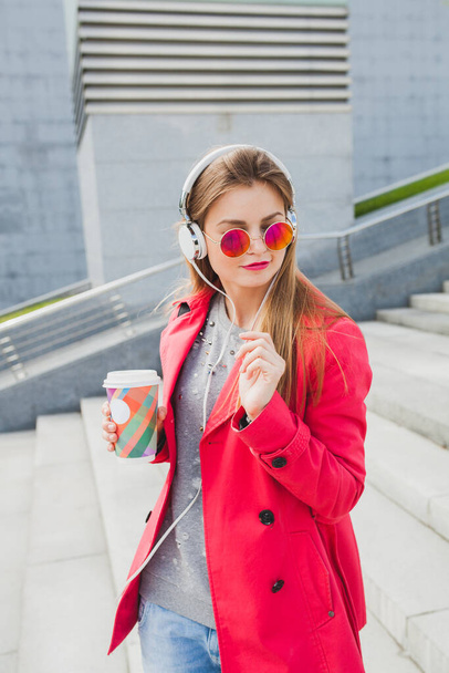 young hipster woman in pink coat, jeans in street with coffee listening to music on headphones, big city urban style trend, wearing sunglasses - Fotografie, Obrázek