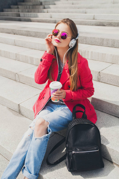 young hipster woman in pink coat, jeans in street with backpack and coffee listening to music on headphones, big city urban style trend, wearing sunglasses - Photo, image