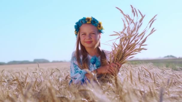 小麦畑のスパイクレットの間で彼女の頭の上に花輪を持つ少女の肖像画。晴れた夏の日に収穫します。滑らかなカメラの動き. - 映像、動画