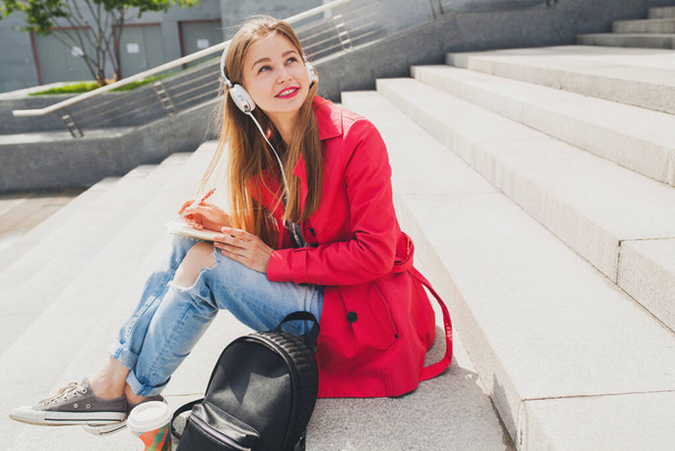 young hipster woman in pink coat, jeans sitting in street with backpack and coffee listening to music on headphones, big city urban style trend, student making notes - Foto, Imagem
