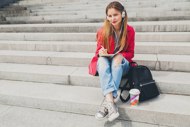 young hipster woman in pink coat, jeans sitting in street with backpack and coffee listening to music on headphones, big city urban style trend, student making notes - Photo, image