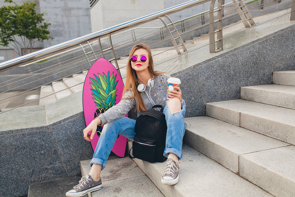 young hipster woman in street with balance board wearing sweater and jeans, spring summer style, student on vacation, urban trend, headphones - Zdjęcie, obraz