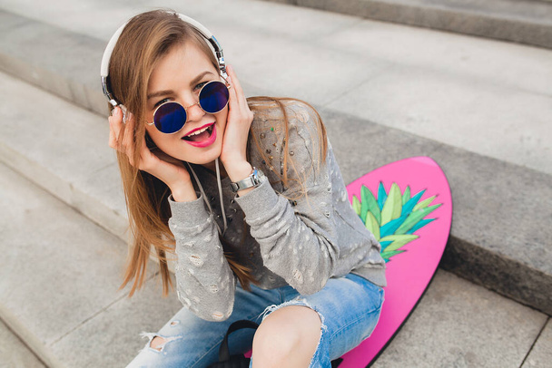 young hipster woman in street with balance board wearing sweater and jeans, spring summer style, student on vacation, urban trend, listening to music on headphones - Foto, imagen