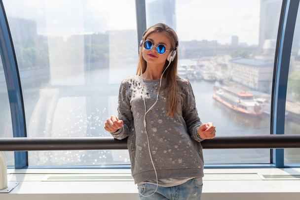 young hipster woman in casual outfit having fun listening to music in headphones, student on vacation, wearing jeans, sweater and sunglasses, urban style sitting at window with city view - Foto, afbeelding