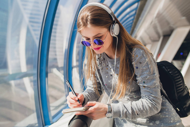 young hipster woman in casual outfit having fun listening to music in headphones, wearing sweater and sunglasses, student making notes - Foto, imagen