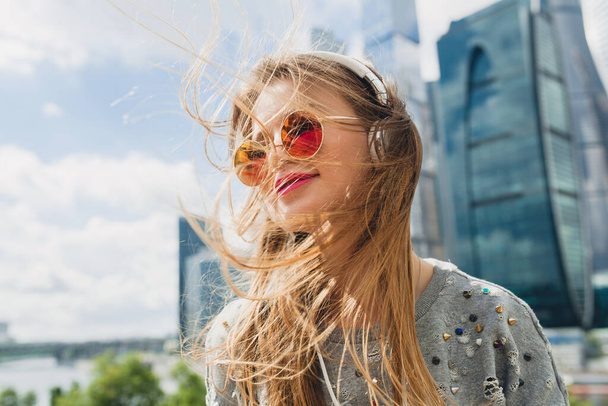 young hipster woman having fun in street listening to music on headphones, wearing pink sunglasses, student on vacation, spring summer urban style, long blond hair - Foto, imagen