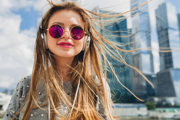 young hipster woman having fun in street listening to music on headphones, wearing pink sunglasses, student on vacation, spring summer urban style, long blond hair - Fotó, kép