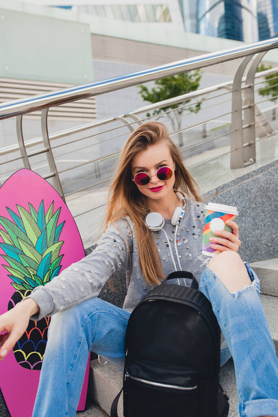 young hipster woman in street with balance board wearing sweater and jeans, spring summer style, student on vacation, urban trend, lazy drinking coffee - Photo, image