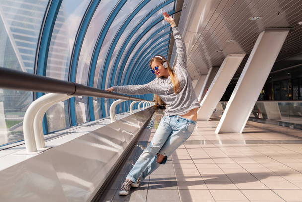 young hipster woman dancing having fun in urban modern building dressed in casual outfit listening to music in headphones, student on vacation, wearing jeans, sweater and sunglasses - 写真・画像