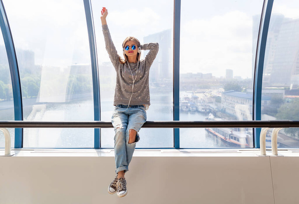 young hipster woman in casual outfit having fun listening to music in headphones, student on vacation, wearing jeans, sweater and sunglasses, urban style sitting at window with city view - Foto, Imagen
