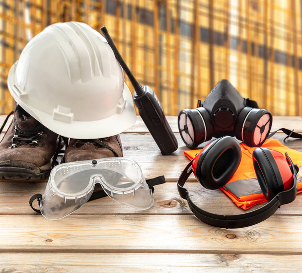 Εξοπλισμός προστασίας της ασφάλειας στην εργασία. Βιομηχανική προστατευτικό εξοπλισμό σε ξύλινο τραπέζι, θολό φόντο εργοτάξιο. Καπέλα, μπότες, ωτοασπίδες, αναπνευστική μάσκα, γυαλιά - Φωτογραφία, εικόνα
