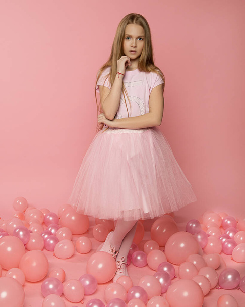 Φωτογραφία ενός όμορφου κοριτσιού 9-10 ετών με ροζ ρούχα σε ροζ φόντο ανάμεσα σε ροζ μπαλόνια. Κομψή φωτογραφία μόδας για κάρτες γενεθλίων, εξώφυλλα, διαφήμιση των διοργανωτών διακοπών - Φωτογραφία, εικόνα